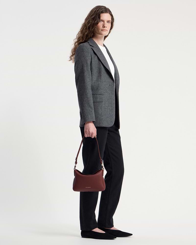 Model holding oxblood Sway shoulder bag by Strathberry while wearing grey blazer.