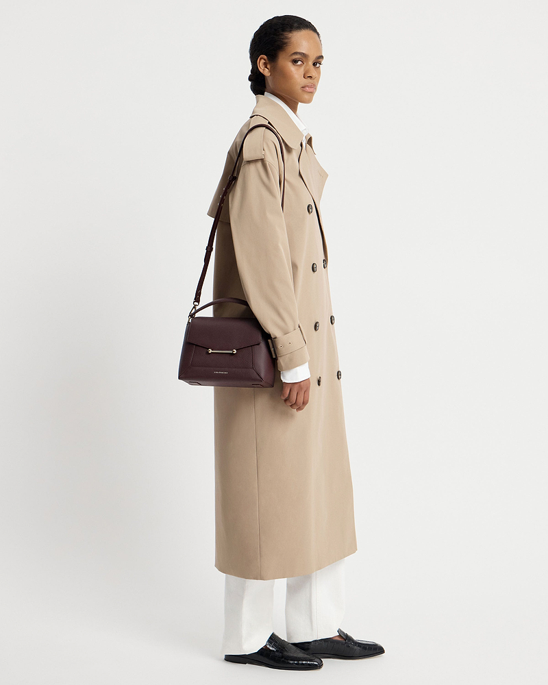 Model in beige trench coat carrying burgundy Strathberry Mosaic bag on shoulder