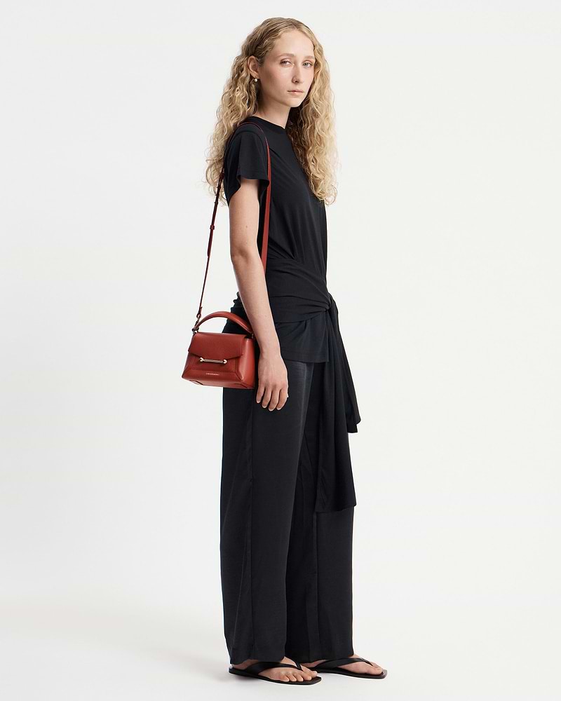 Model in black outfit carrying small red top-handle bag on her shoulder.