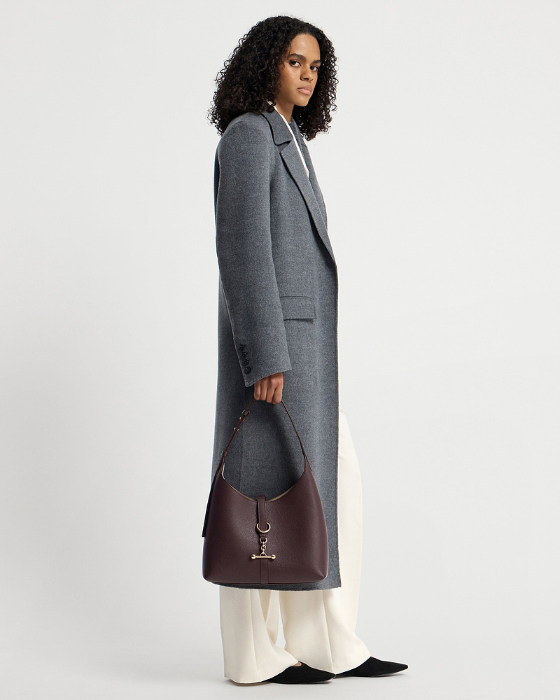 Model holding burgundy hobo bag with gold hardware, wearing grey coat and cream trousers.