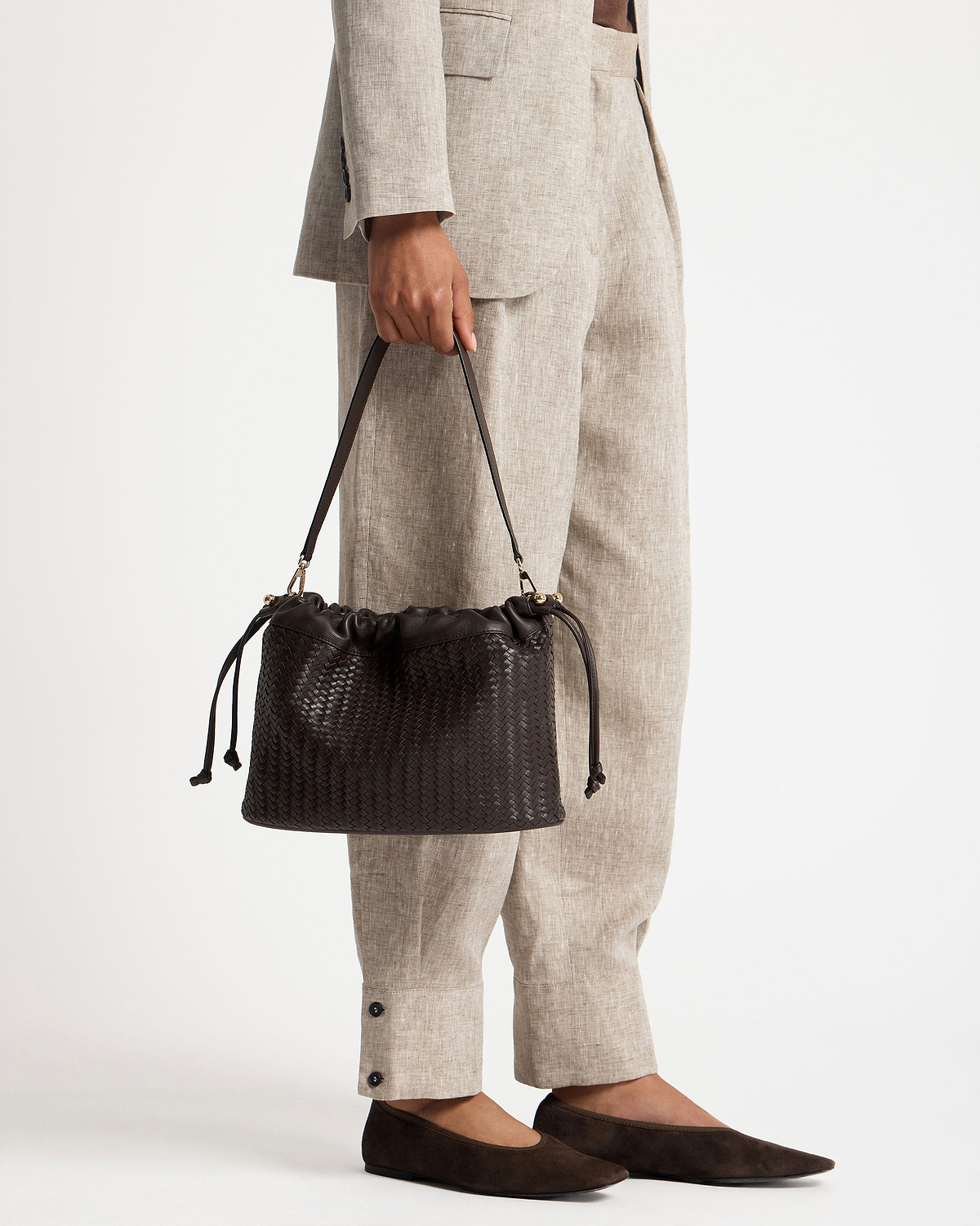 Model holding chocolate woven leather drawstring bag with beige suit trousers and flats