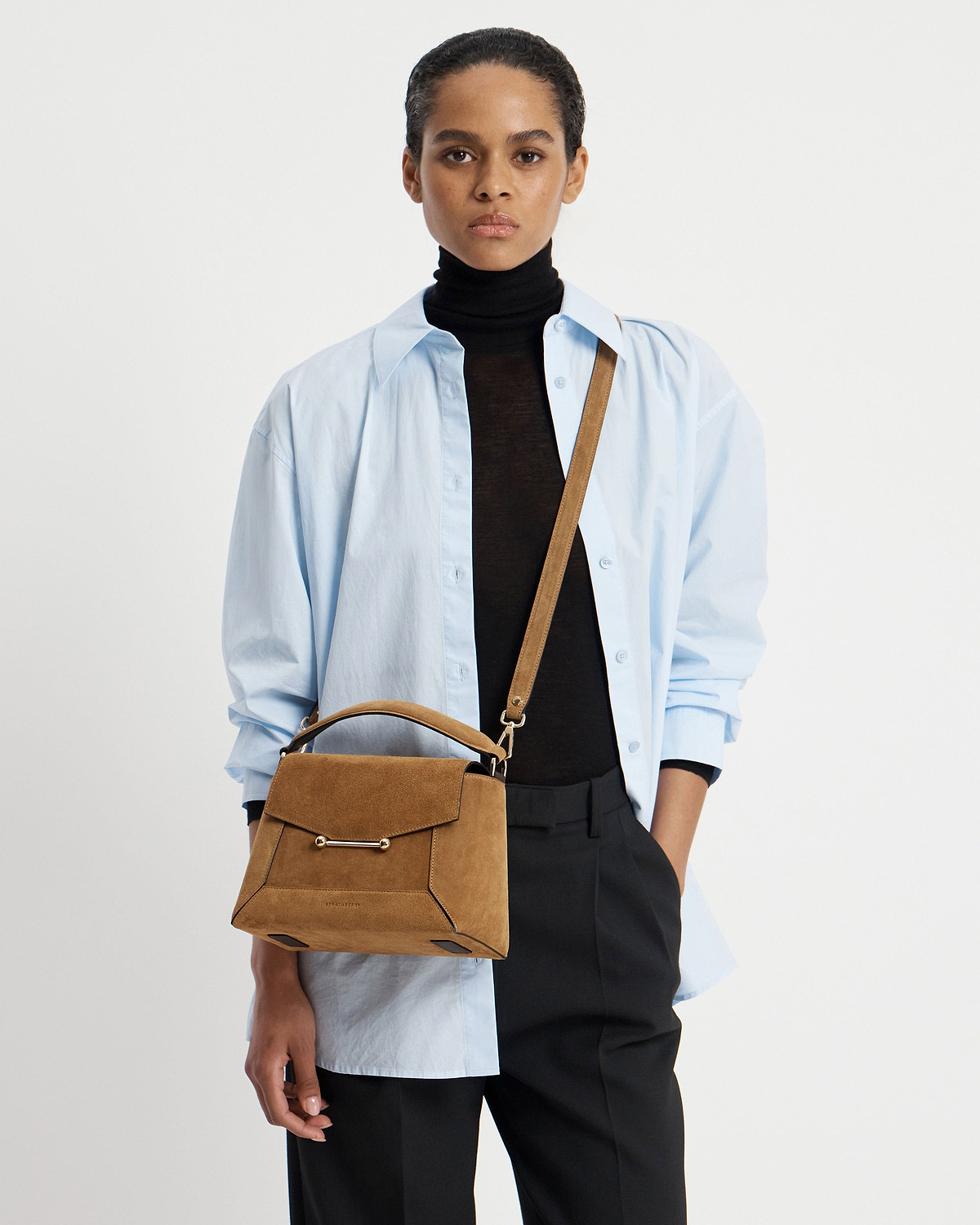 Model wearing light blue shirt and black roll-neck carrying toffee suede mosaic bag.