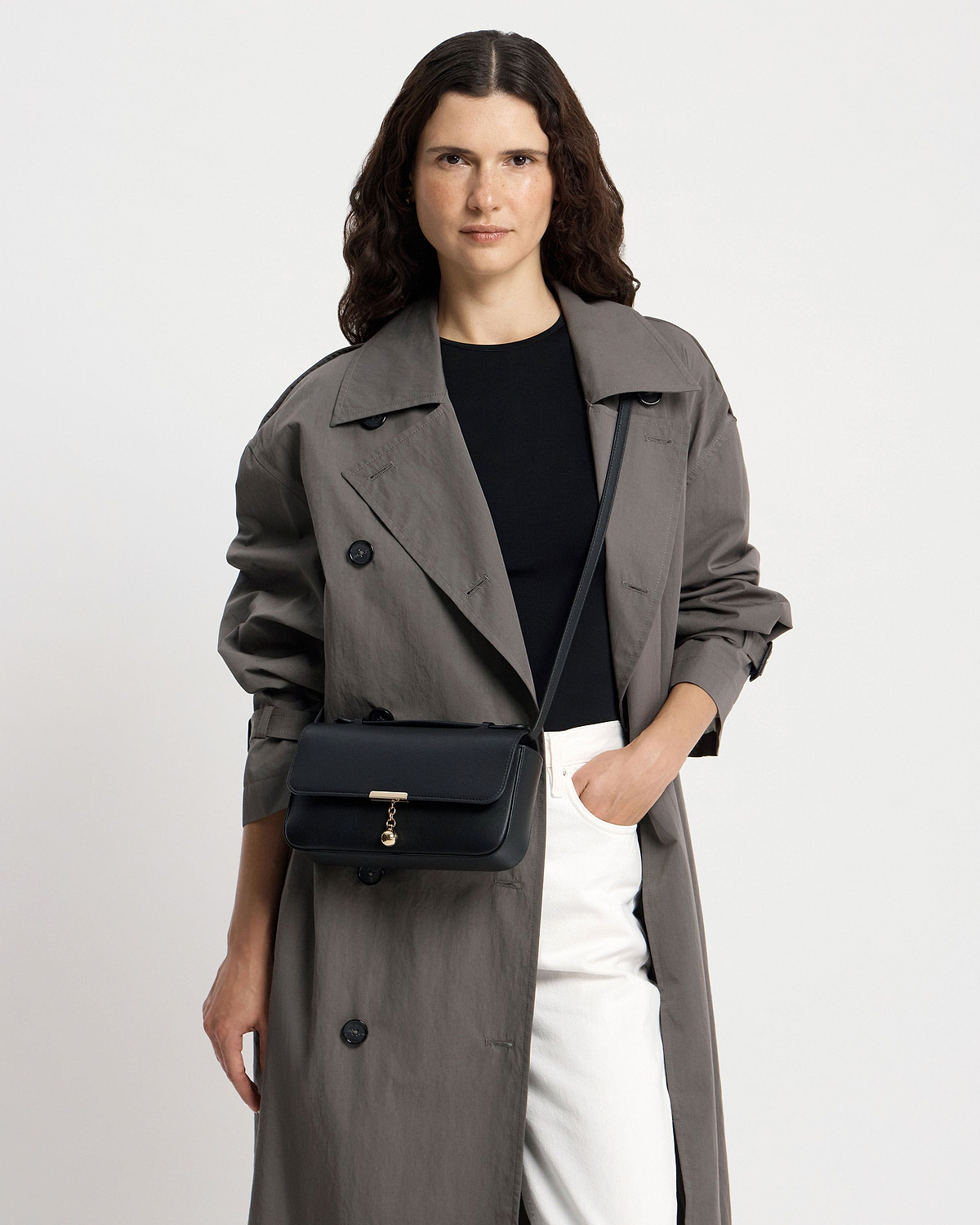 Woman wearing black clutch bag with gold clasp, crossbody strap and grey trench.