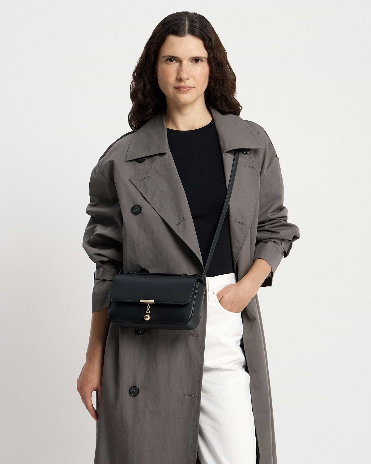 Woman wearing black clutch bag with gold clasp, crossbody strap and grey trench.