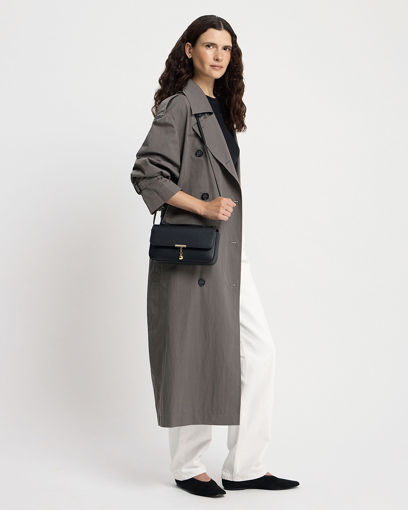 Woman wearing a trench coat carrying a black Nocturn clutch with gold clasp.