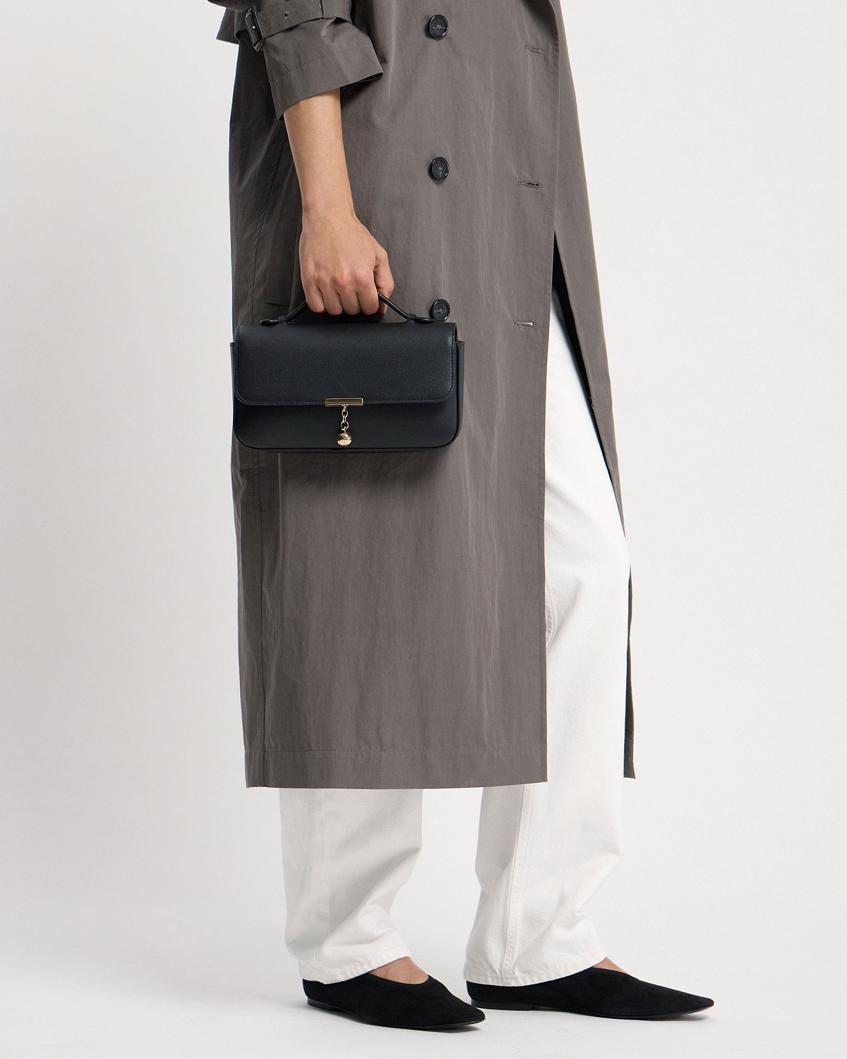 Hand holding black clutch with top handle and gold charm, wearing grey coat.