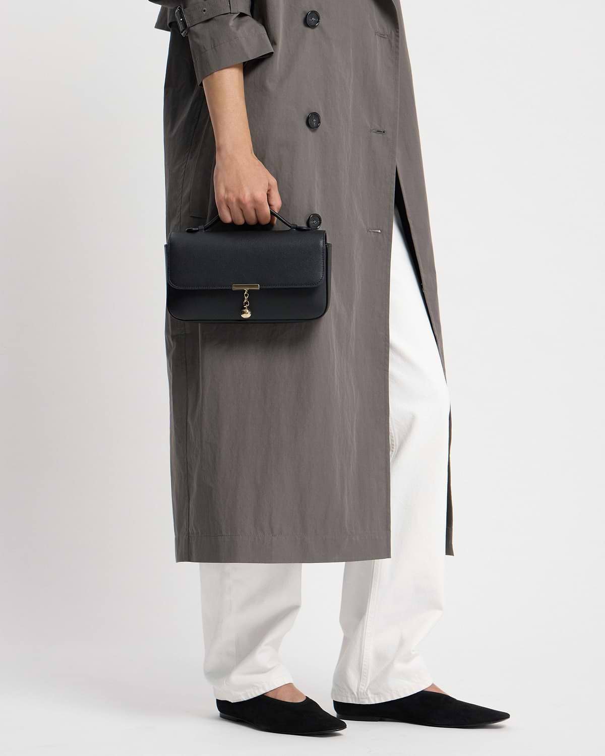 Hand holding black clutch with top handle and gold charm, wearing grey coat.