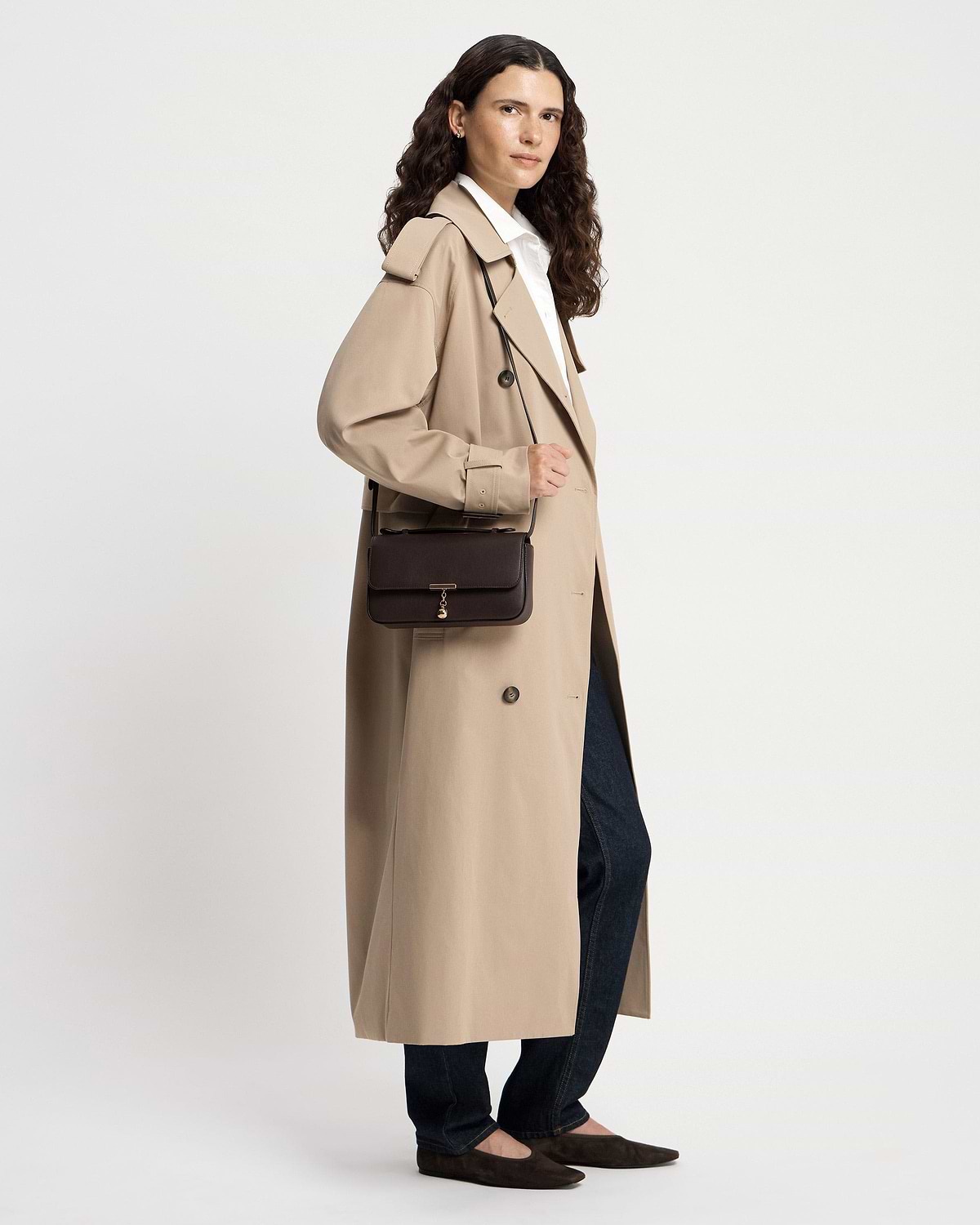 Woman in a beige trench coat holding an espresso leather clutch bag.