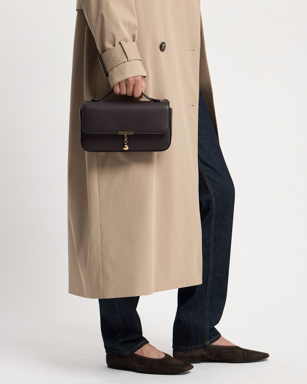 Hand holding espresso leather clutch with top handle and gold charm.