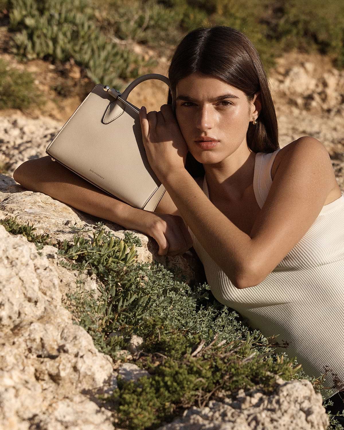 Model holding Strathberry oat mini tote against her shoulder, rocky background.