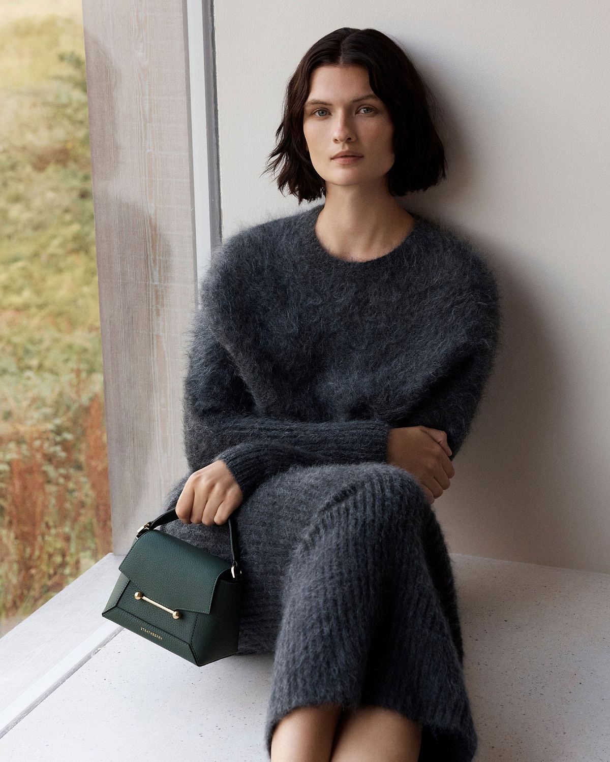 Model seated by window wearing grey fuzzy dress holding bottle green Strathberry Mosaic Nano bag.