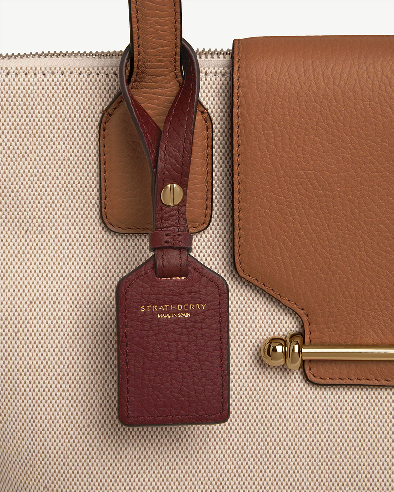 Oxblood leather tag with Strathberry logo attached to beige canvas bag trim and gold hardware.