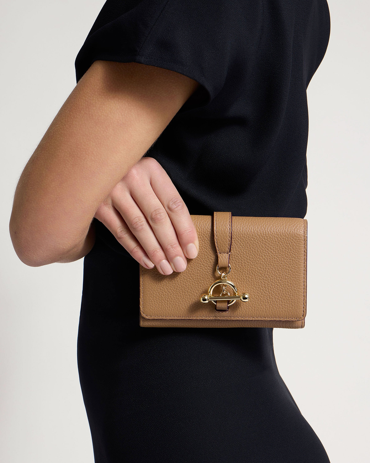 Caramel textured leather trifold wallet held at model's hip, gold bar clasp visible.