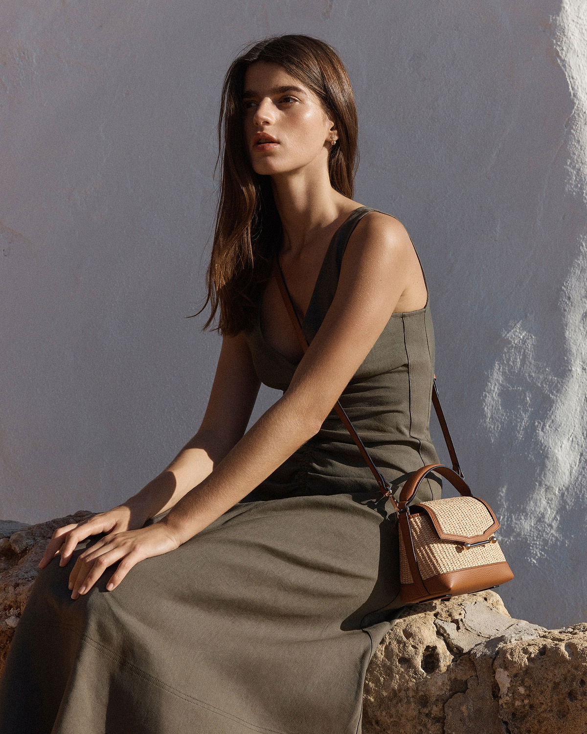 Woman seated in olive dress wearing small tan leather and natural raffia crossbody bag.