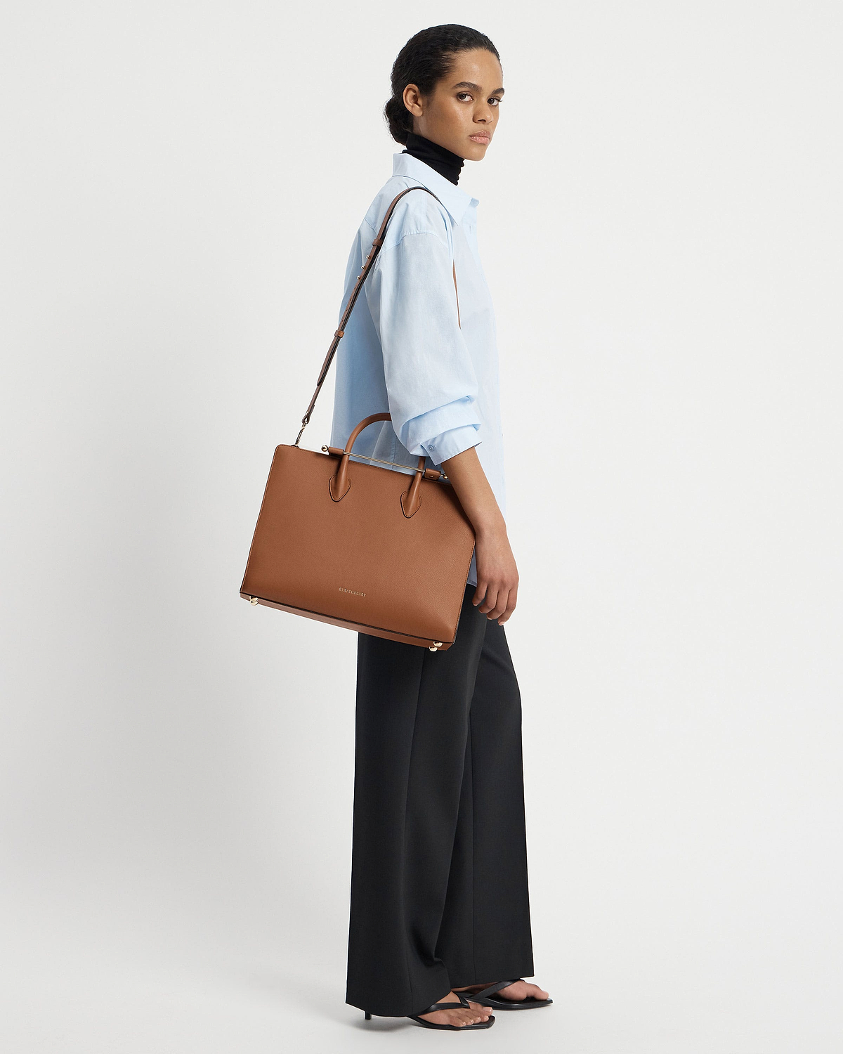 Model wearing a light blue shirt carrying a Strathberry chestnut leather tote.