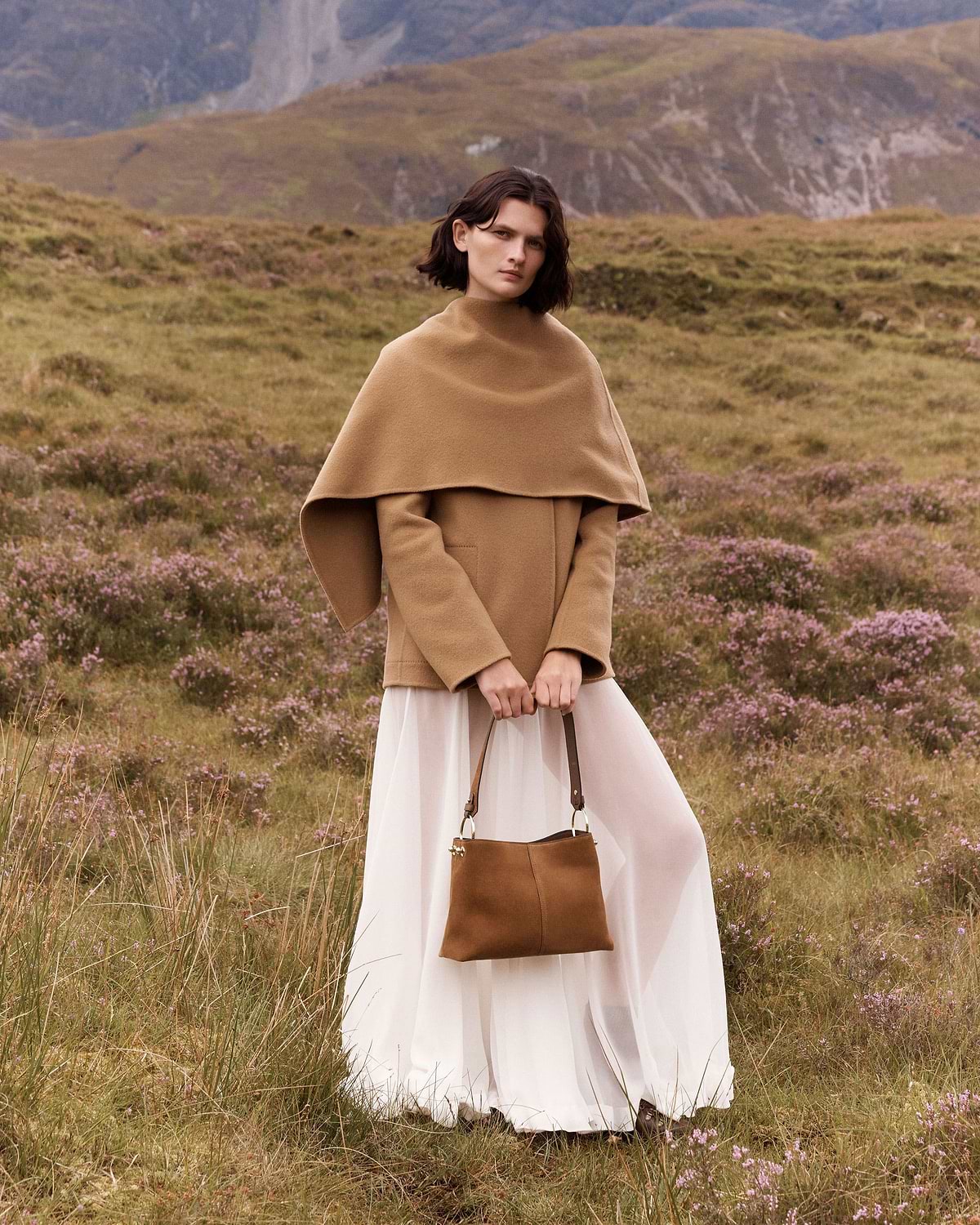 Woman wearing tan coat holding tan suede hobo bag in grassy hillside setting.