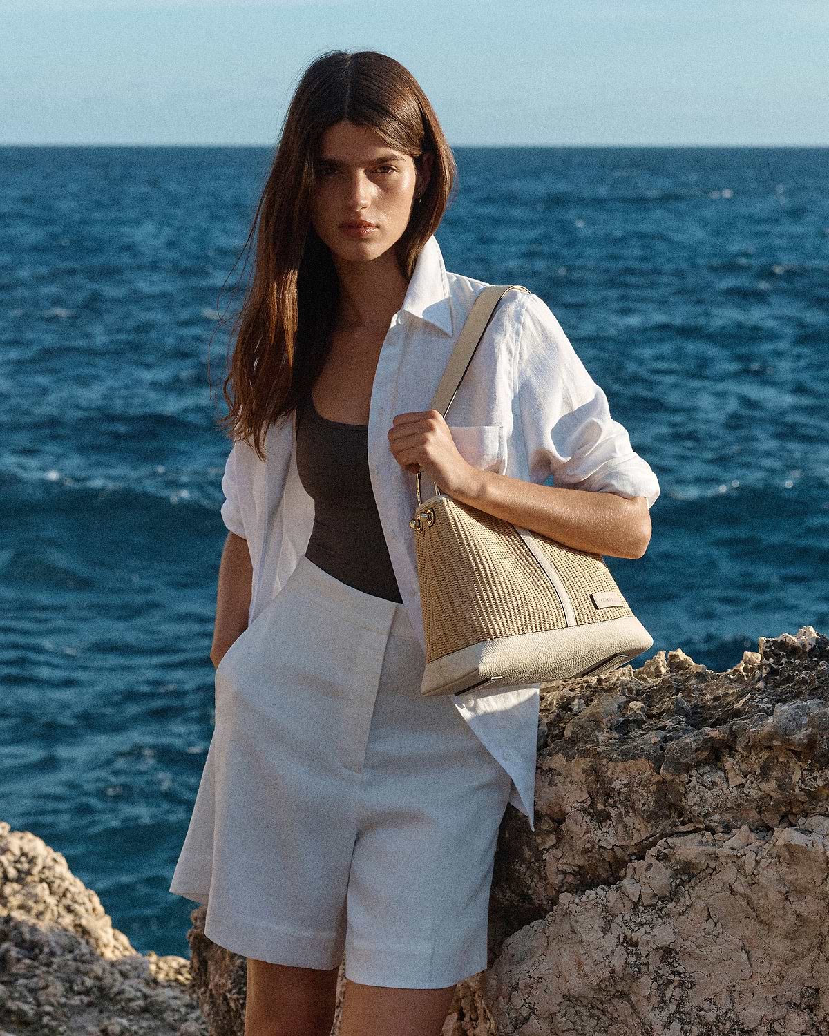 Woman in white shirt holding a vanilla natural raffia hobo bag by the sea.