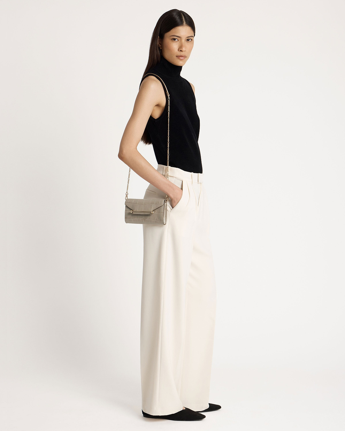 Woman wearing a black sleeveless top and cream trousers, shoulder croc chain wallet.
