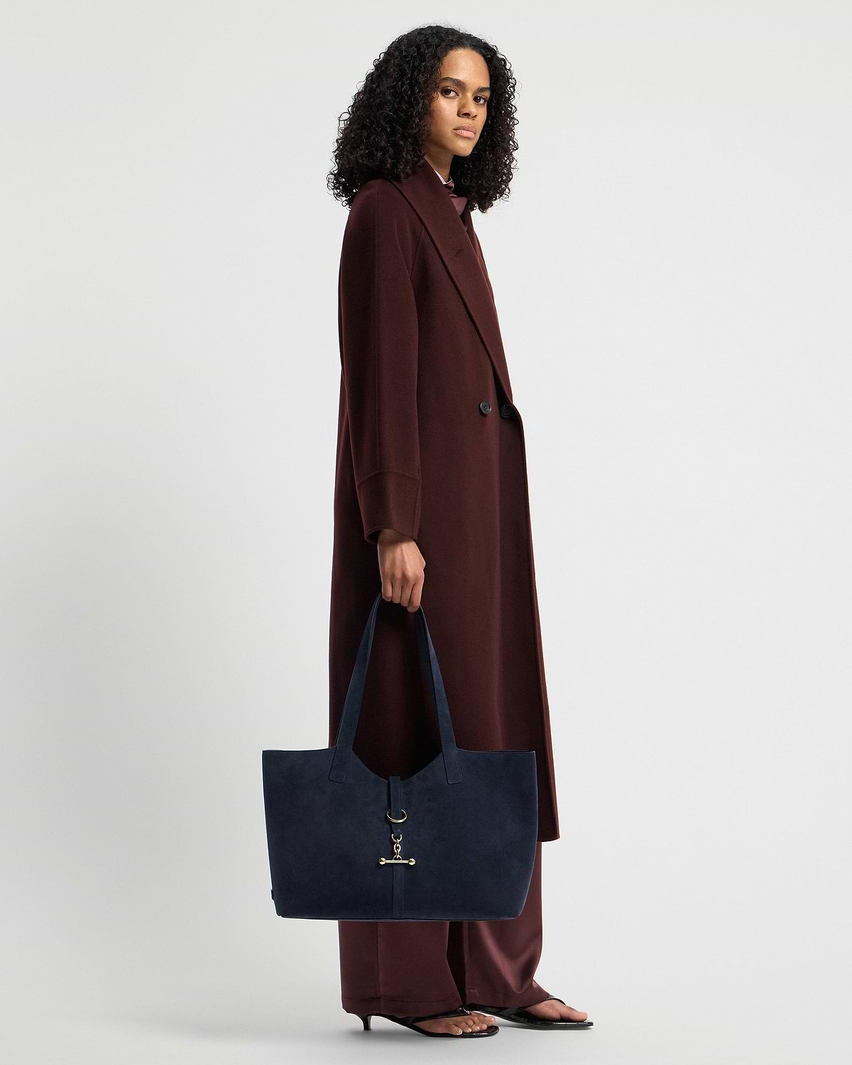 Model holding navy suede kite tote with gold hardware at her side, wearing long coat.
