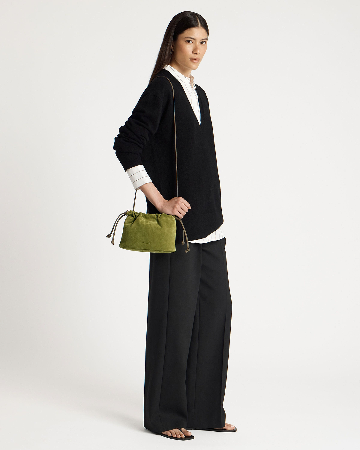 Model wearing black V-neck sweater holds lime suede drawstring bag at her hip.