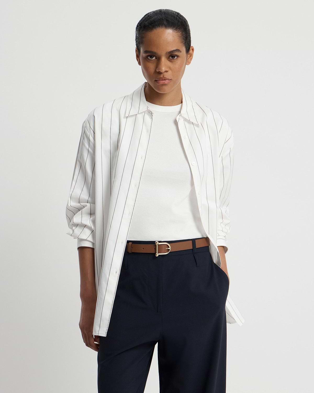 Model wearing white striped shirt, white T-shirt, navy trousers and hazelnut belt with gold buckle.