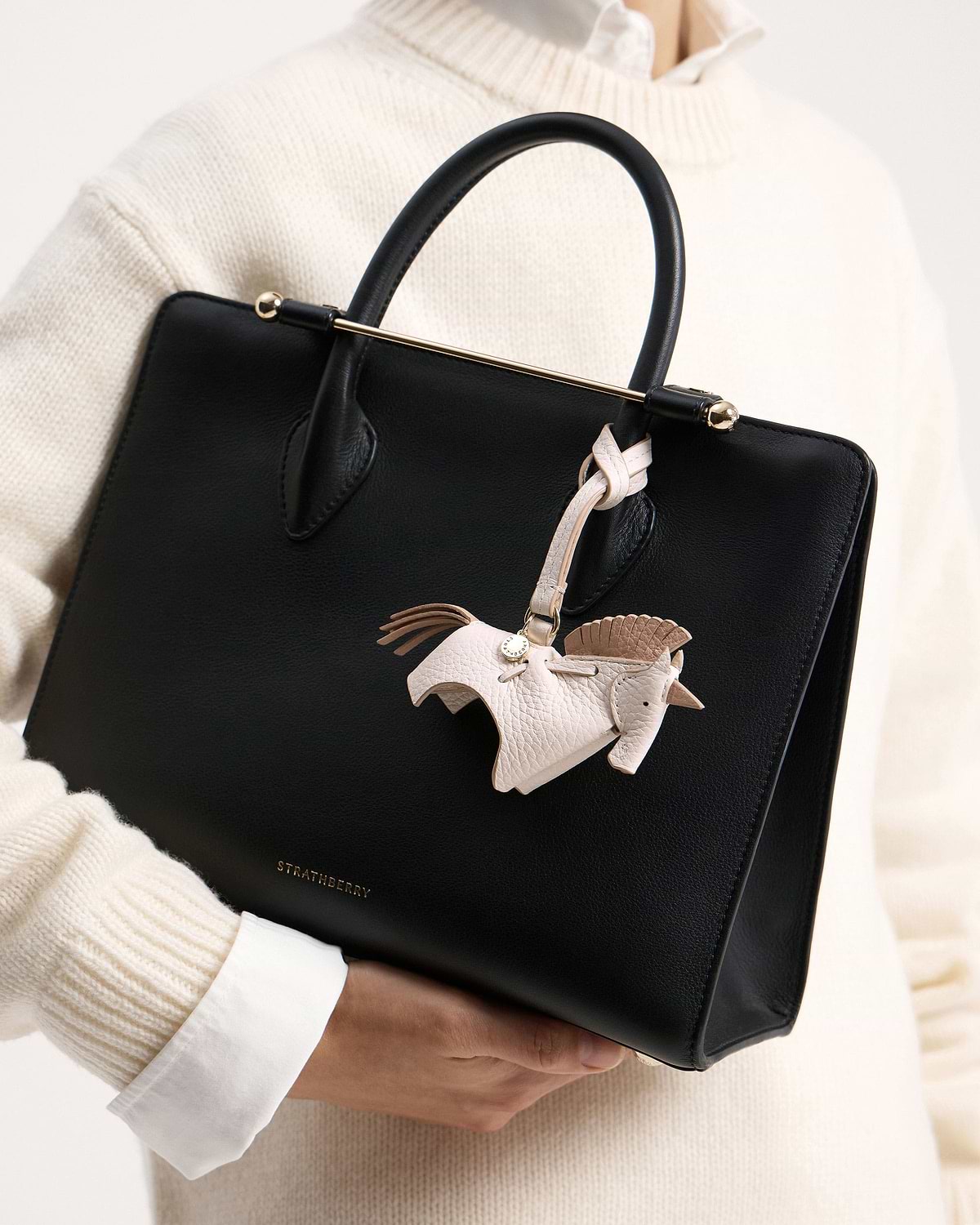 White peony unicorn bag charm on black Strathberry leather bag held by model.