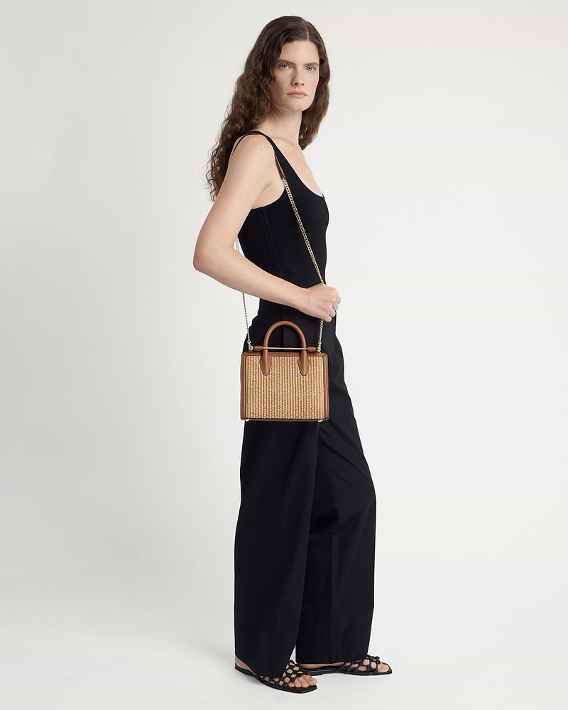 Model in black holds tan mini tote in natural raffia with gold chain strap.