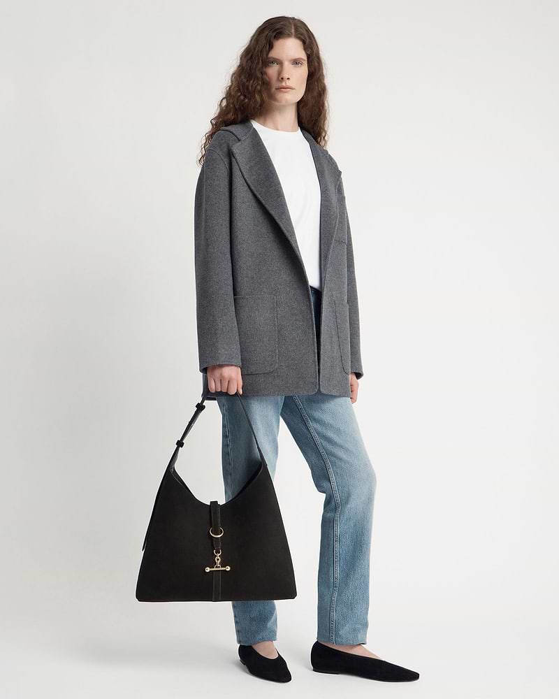 Model wearing grey coat and jeans holding black suede hobo bag with gold hardware.