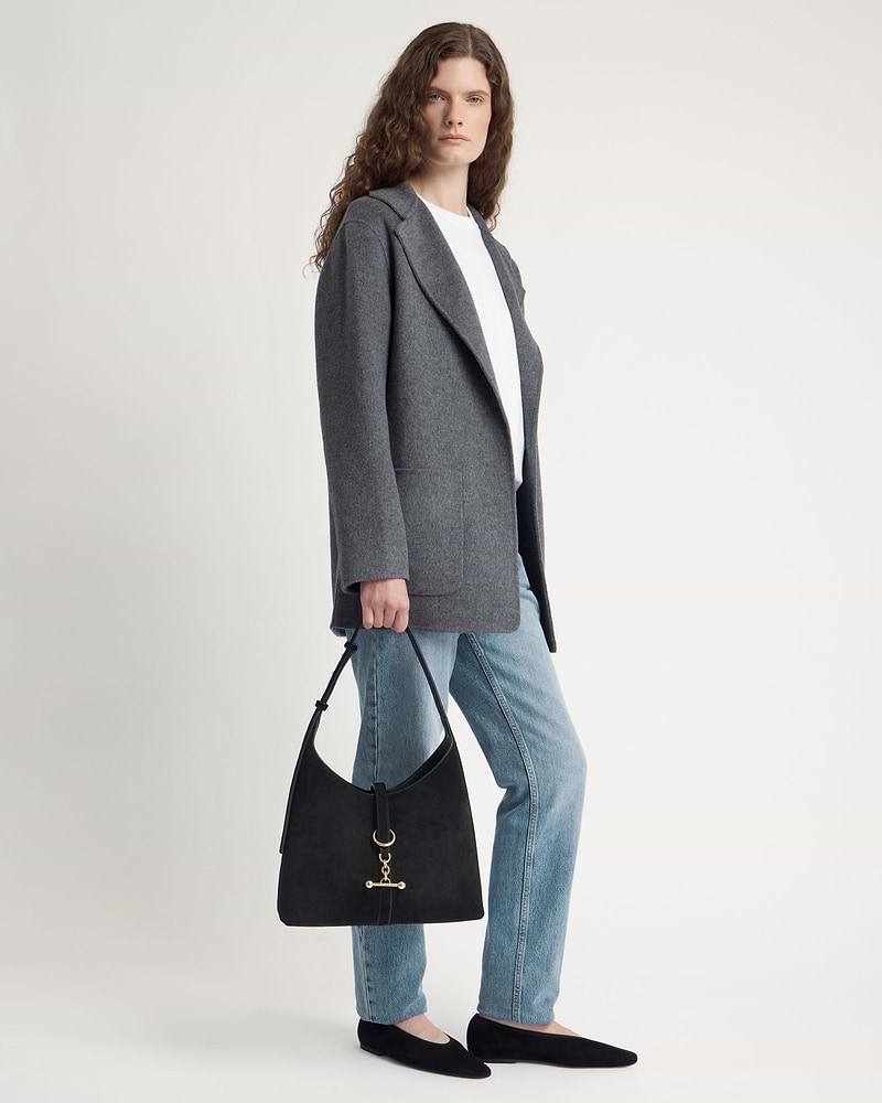 Model in grey coat and jeans holding black suede hobo bag with gold hardware.