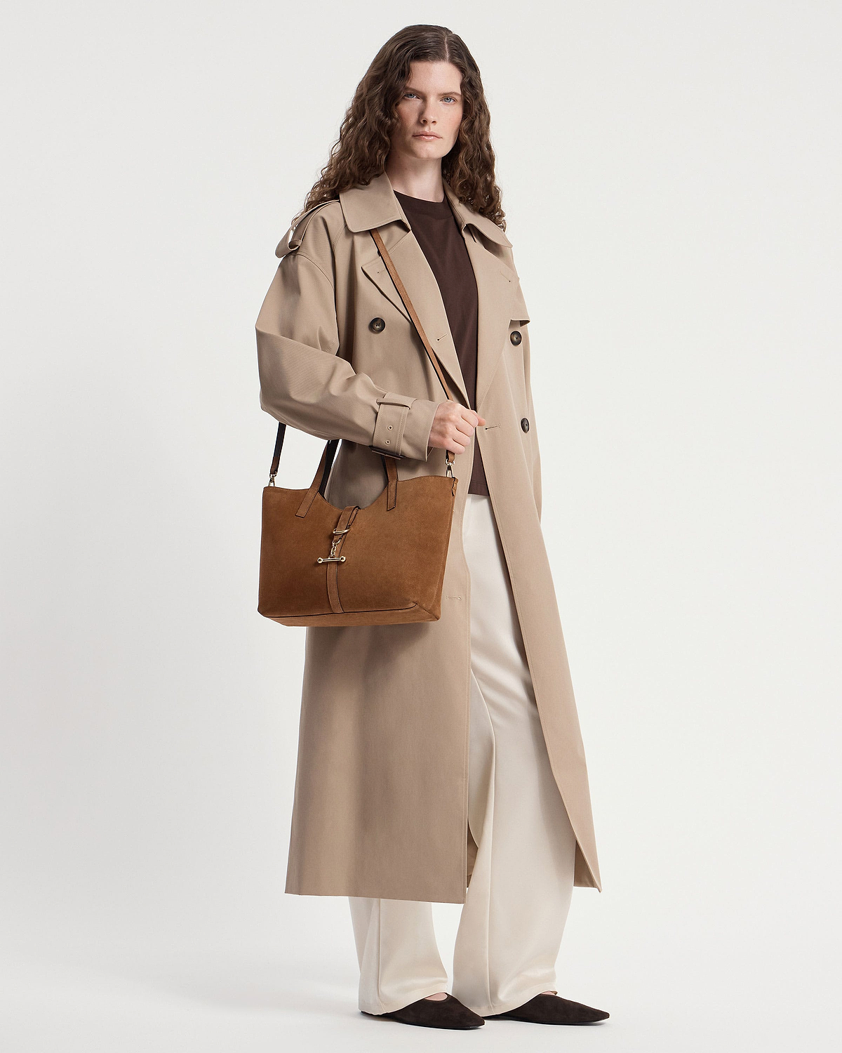 Model in a beige trench coat carrying a tan suede tote bag at their side.