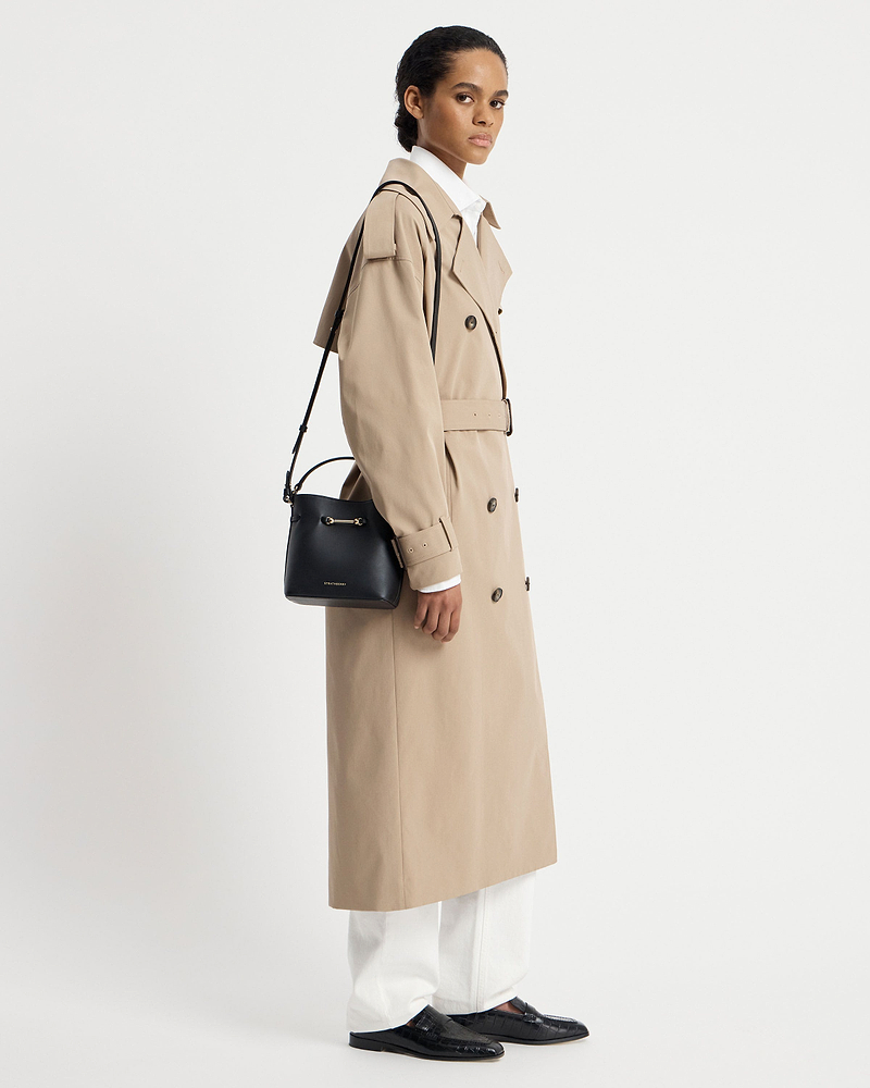 Model wearing a beige trench coat carrying a black Strathberry Corda Bucket Mini shoulder bag