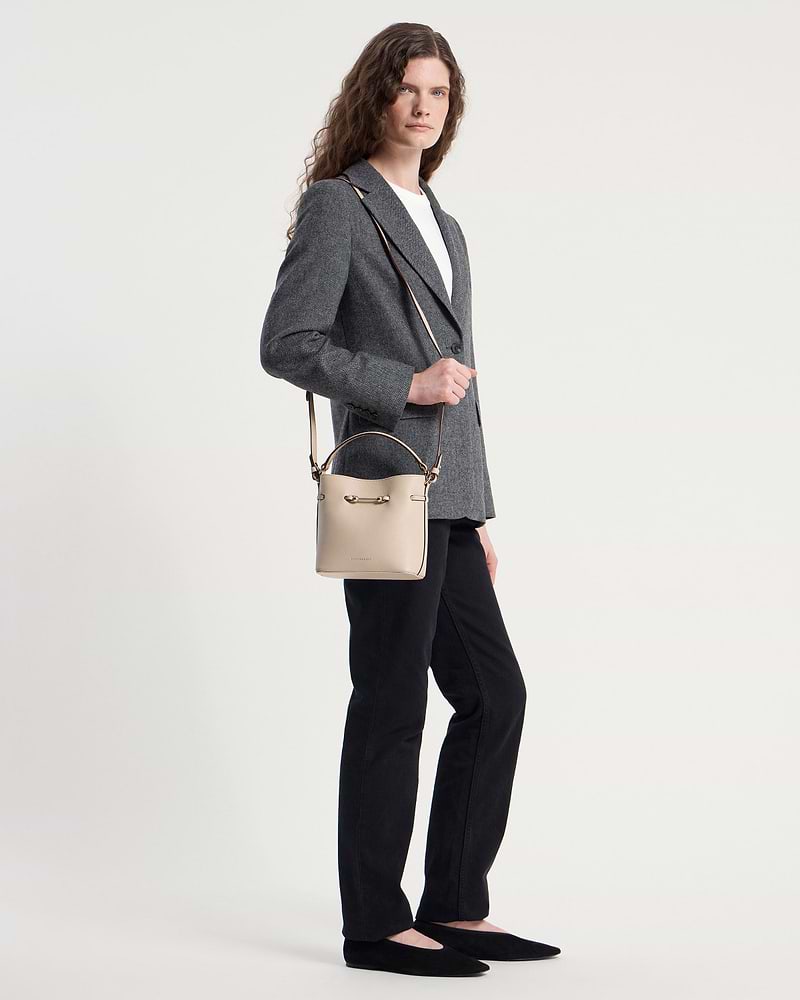 Model wearing a grey blazer holding an oat-coloured mini bucket bag with a shoulder strap.