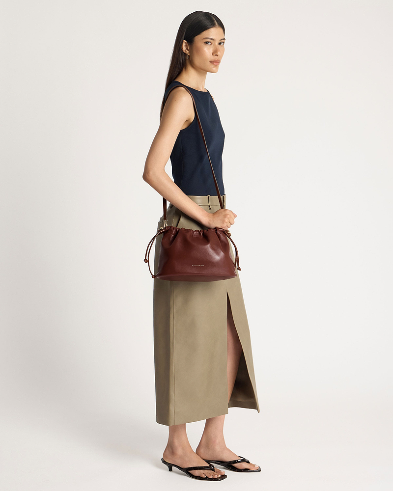 Model wearing a navy top and khaki skirt, carrying a Strathberry walnut drawstring bag.