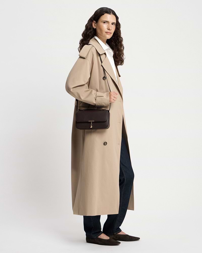 Model in trench coat wearing espresso Nocturn clutch with gold charm and shoulder strap.