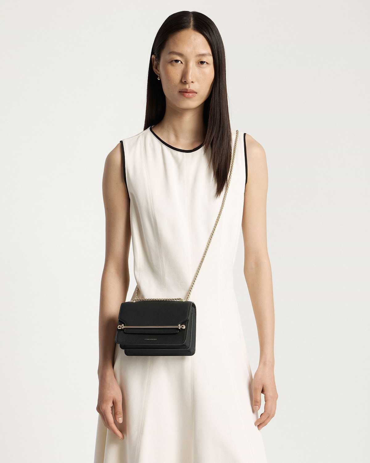 Model in sleeveless white dress wearing black East/West Mini crossbody bag with gold chain