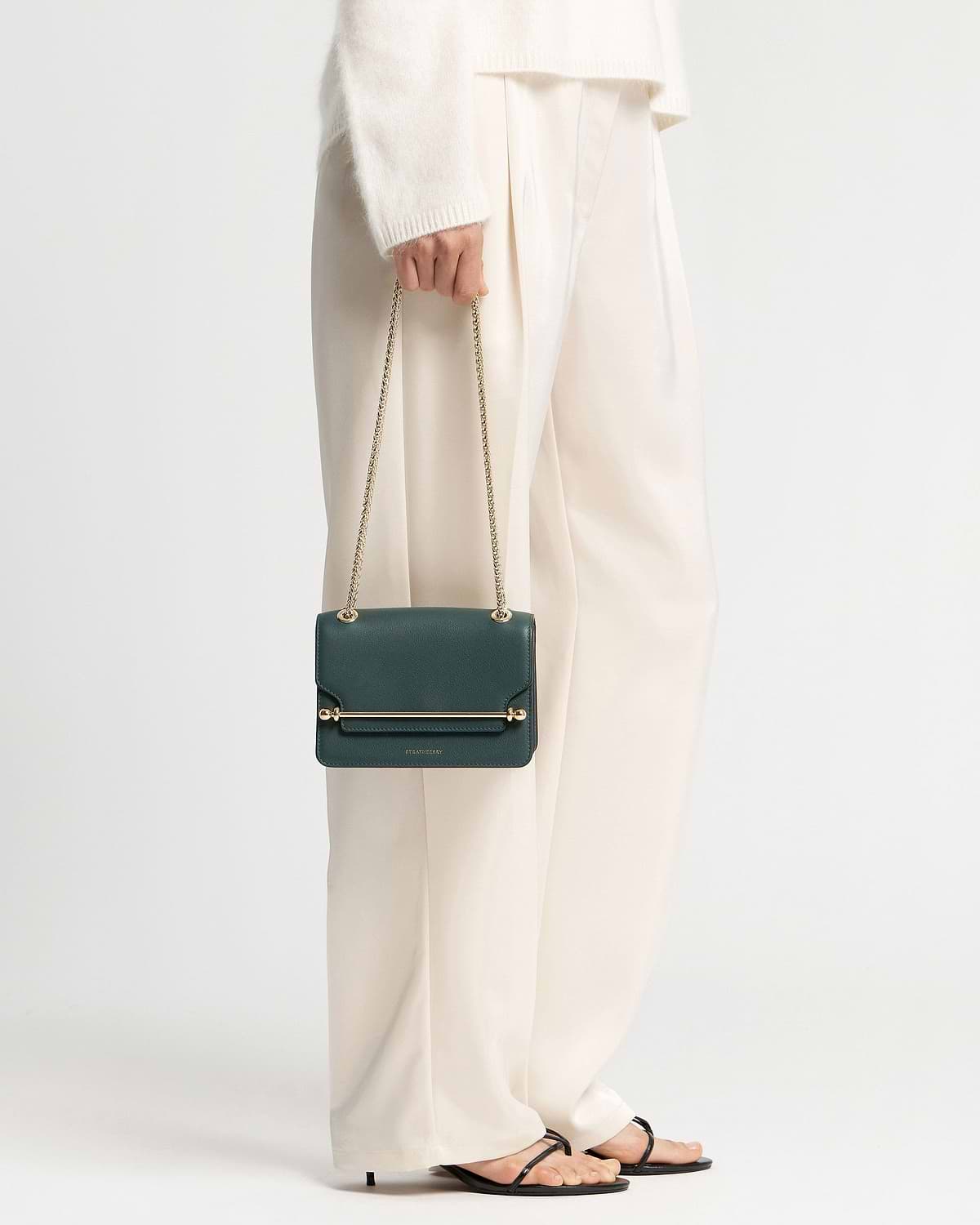 Model holding bottle green Strathberry East/West Mini bag with gold chain strap.