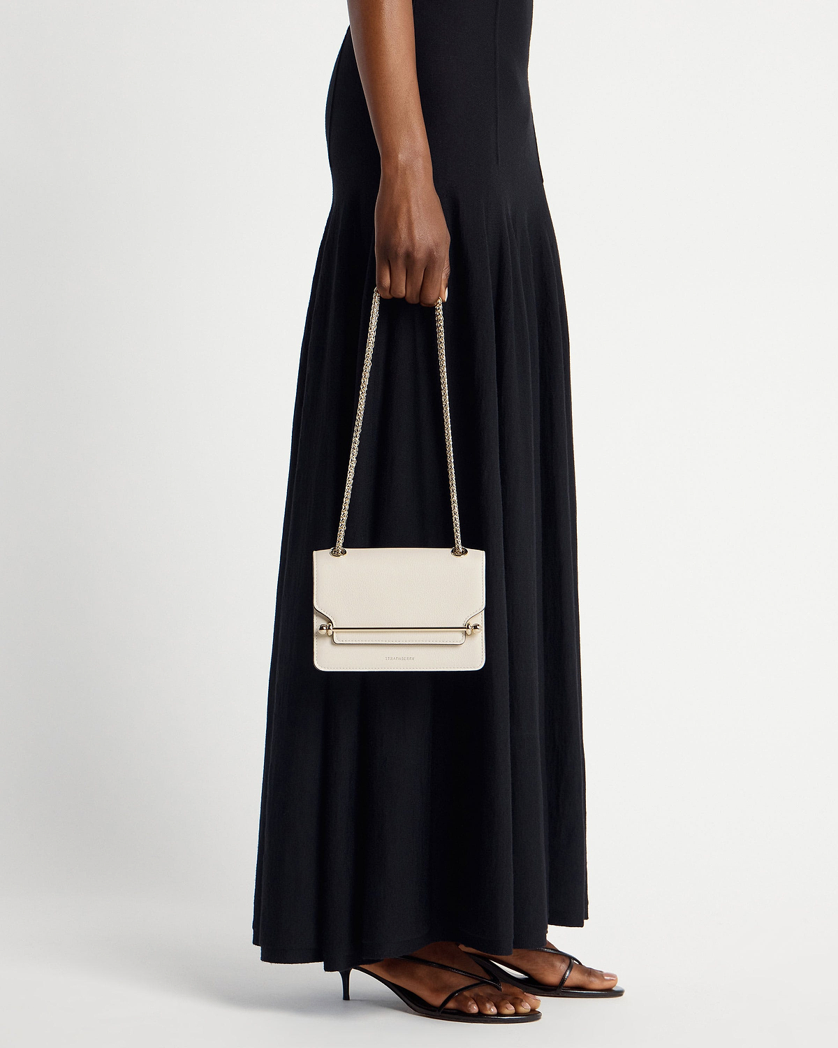 Model holding vanilla East/West Mini bag by Strathberry with chain strap.