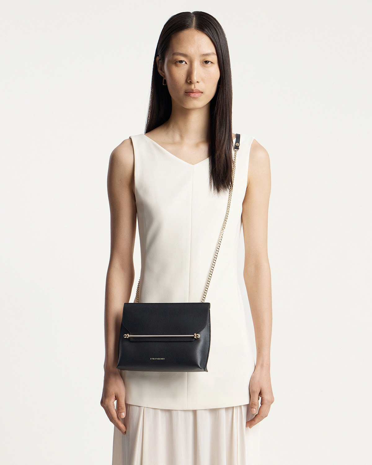 Woman in white dress wearing a black Strathberry crossbody bag with gold hardware