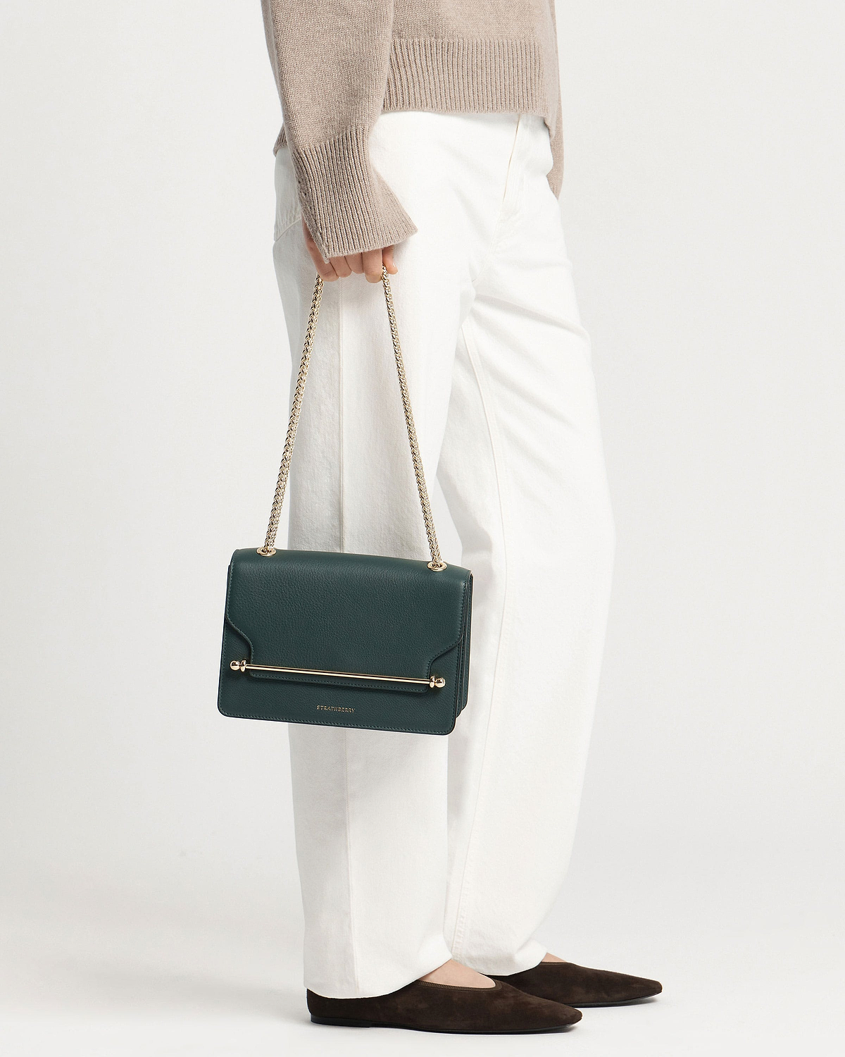 Model holding bottle-green Strathberry East/West bag with gold chain strap.