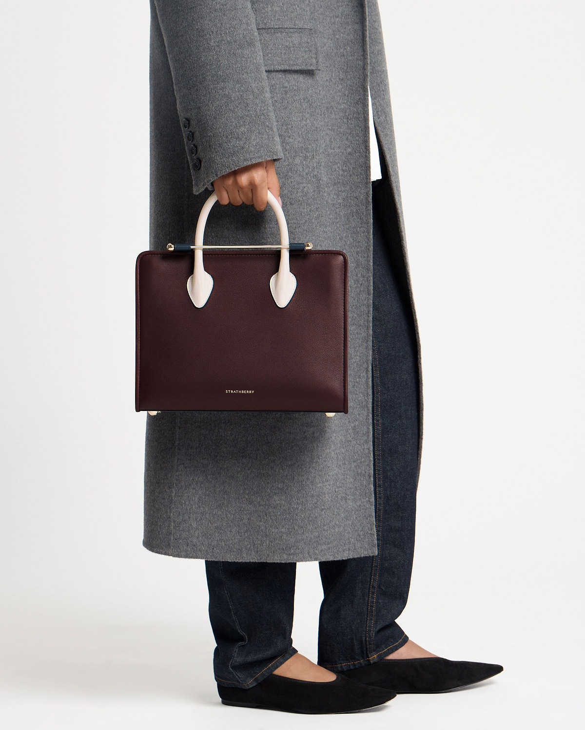 Model cropped below waist holding Strathberry midi tote in burgundy with vanilla and navy handles.