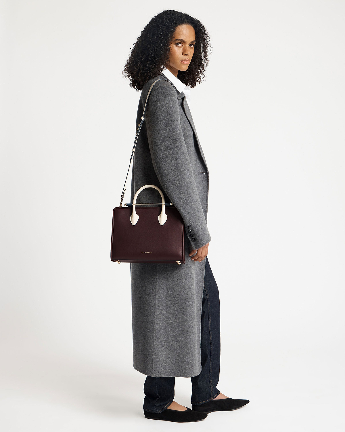Model wearing a grey coat holding a Strathberry midi tote in burgundy, navy and vanilla