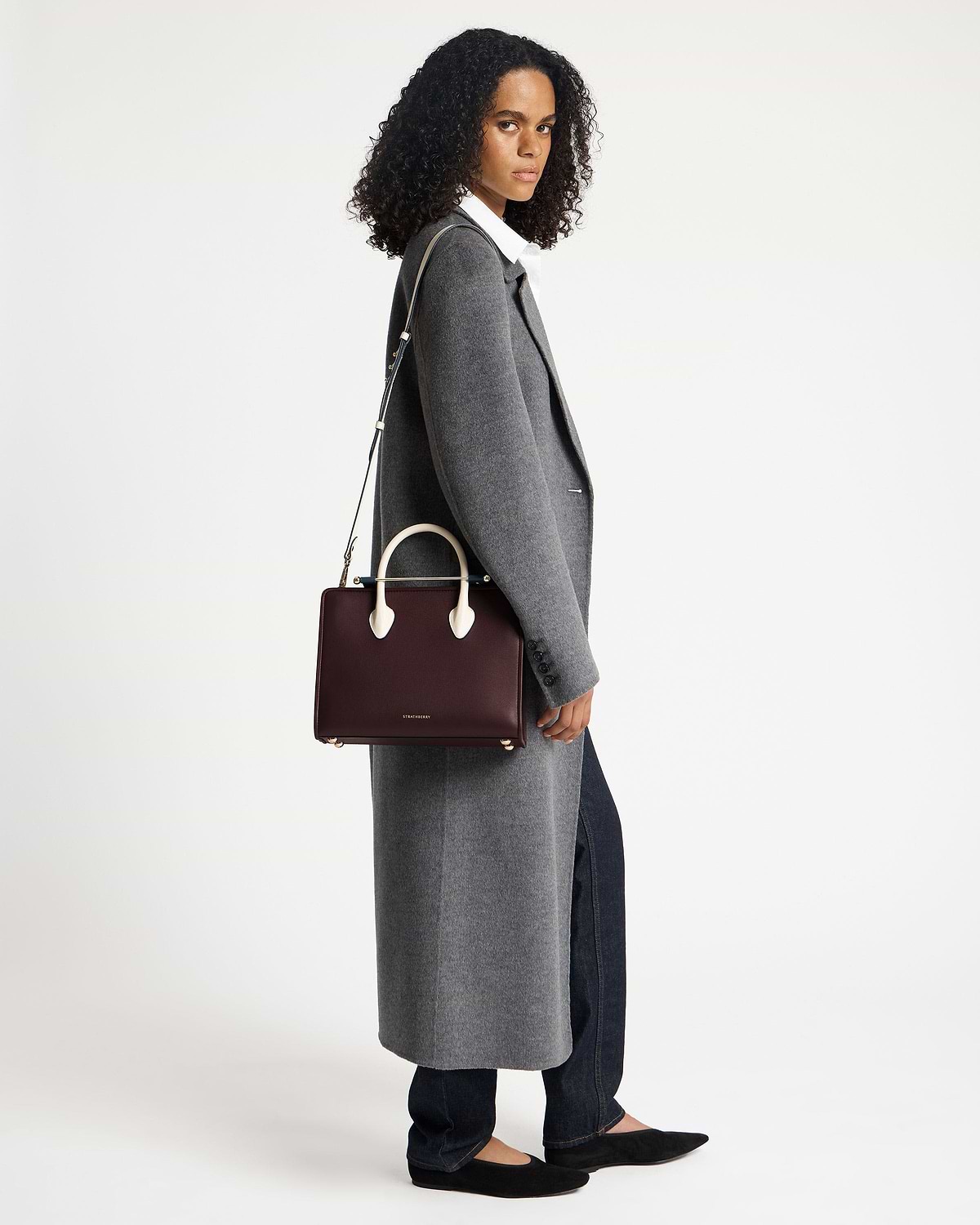 Model wearing a grey coat holding a Strathberry midi tote in burgundy, navy and vanilla
