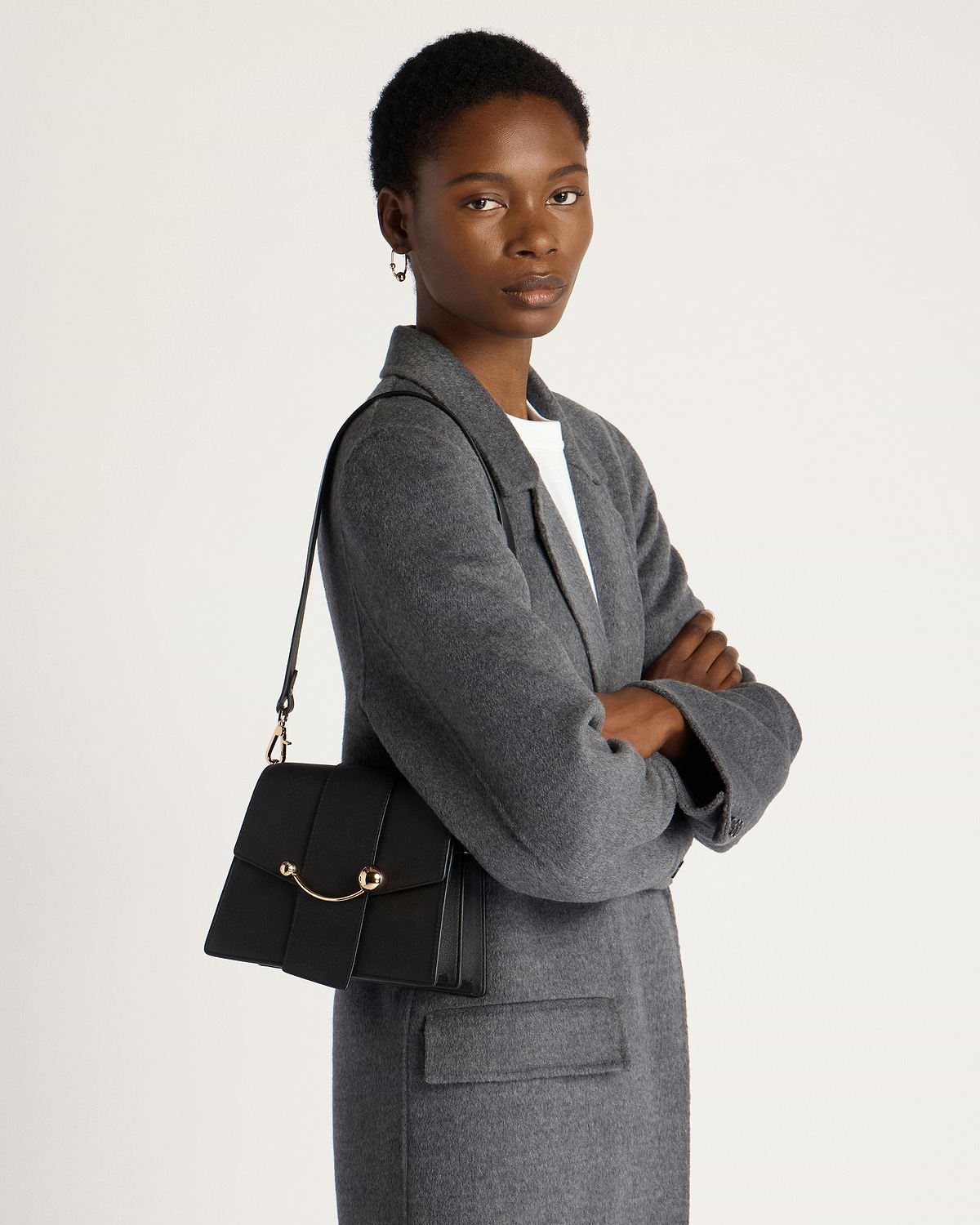 Model wearing grey coat carrying black Box Crescent shoulder bag with gold hardware