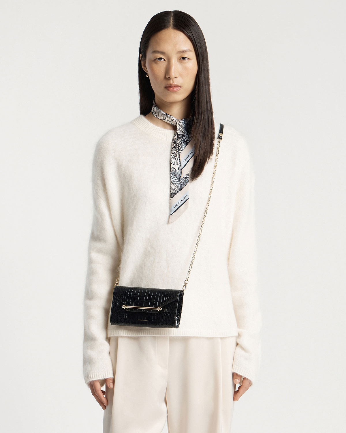 Woman wearing black croc chain wallet with gold bar detail, plus Strathberry neck scarf.