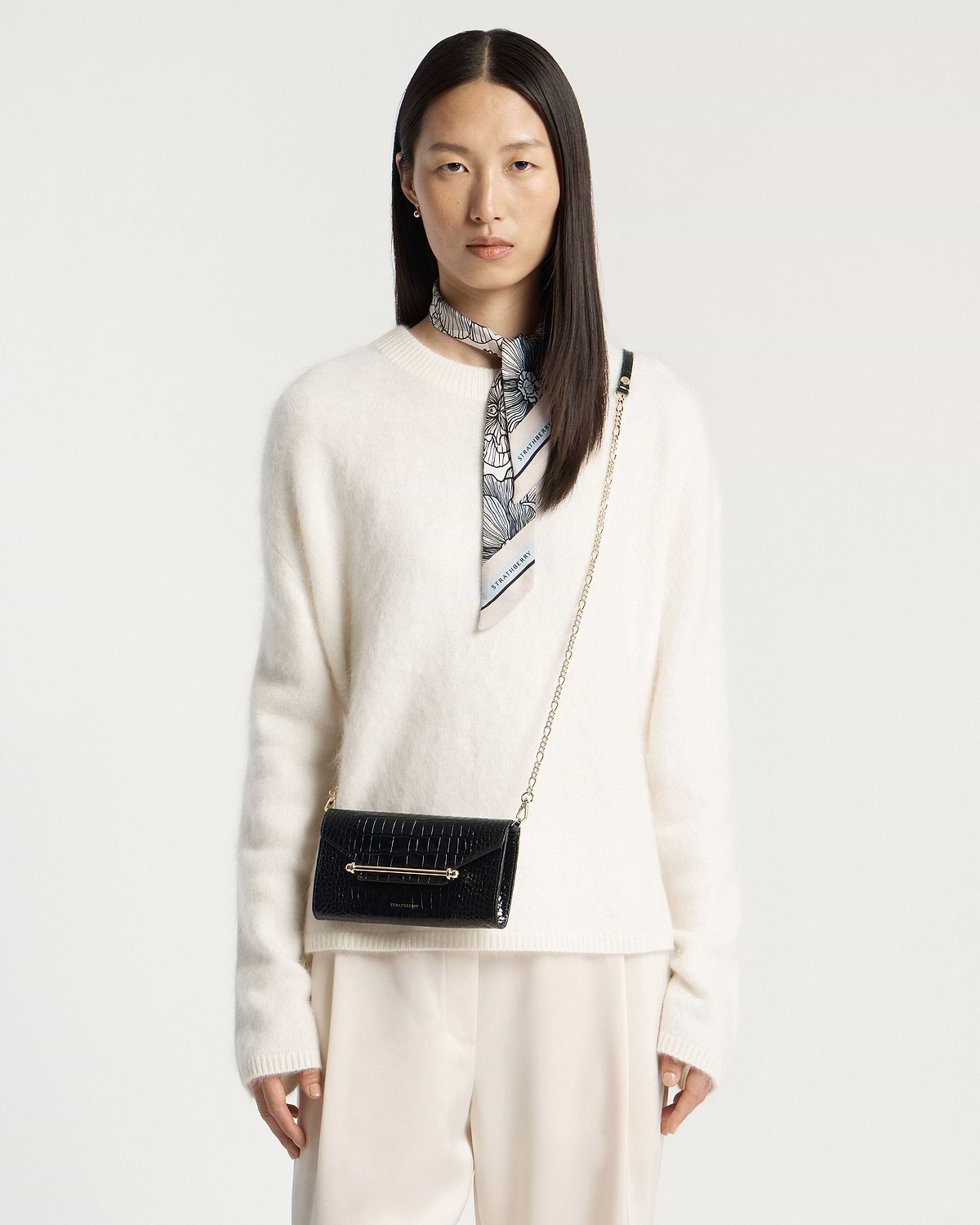 Woman wearing black croc chain wallet with gold bar detail, plus Strathberry neck scarf.