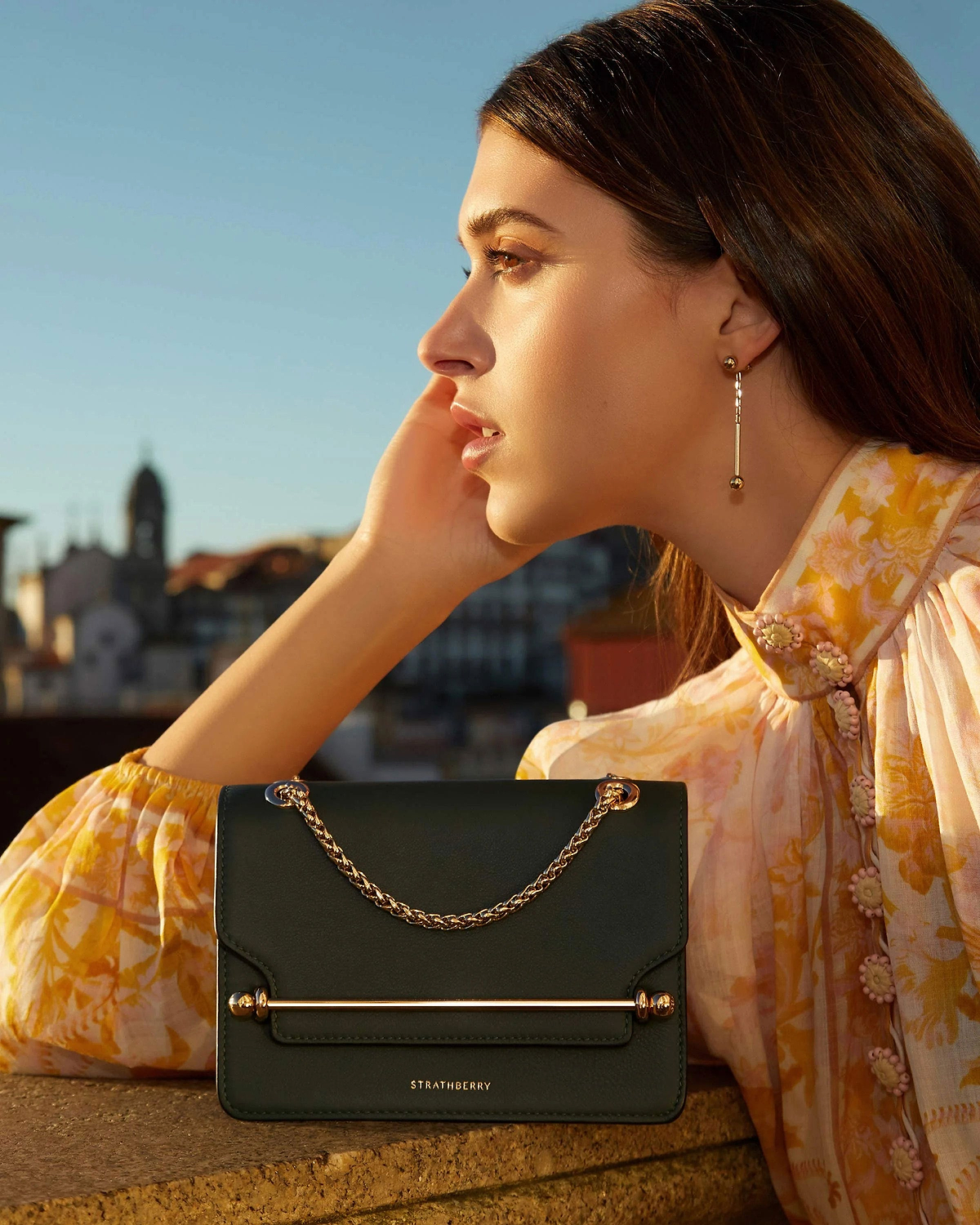 Profile of woman wearing long 22-carat gold bar drop earrings beside Strathberry handbag.