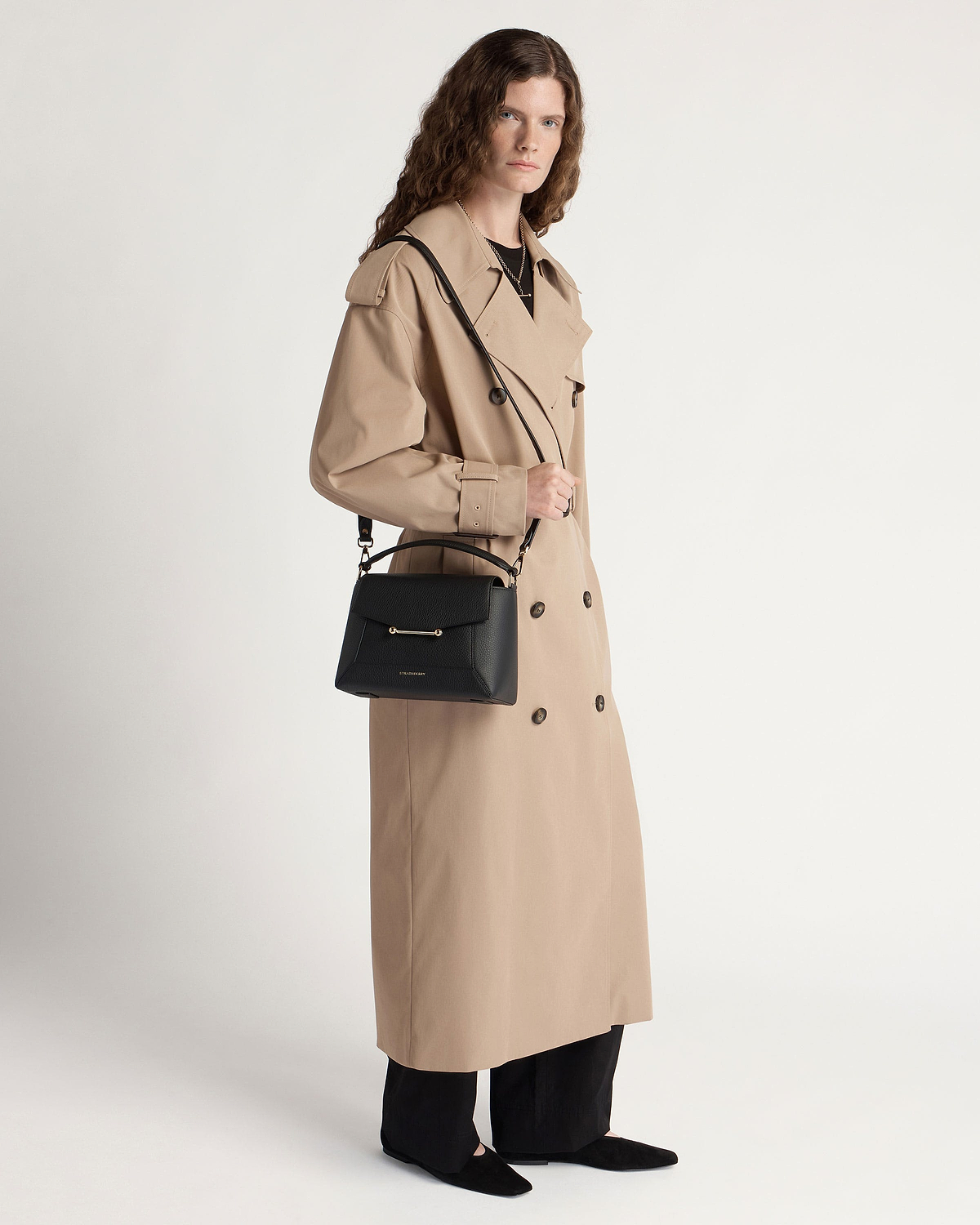 Model in a beige trench coat holding a black mosaic bag with shoulder strap.
