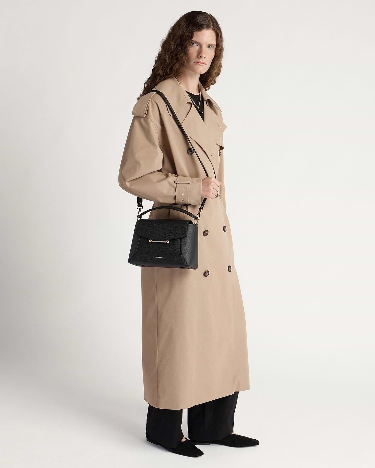 Model in a beige trench coat holding a black mosaic bag with shoulder strap.