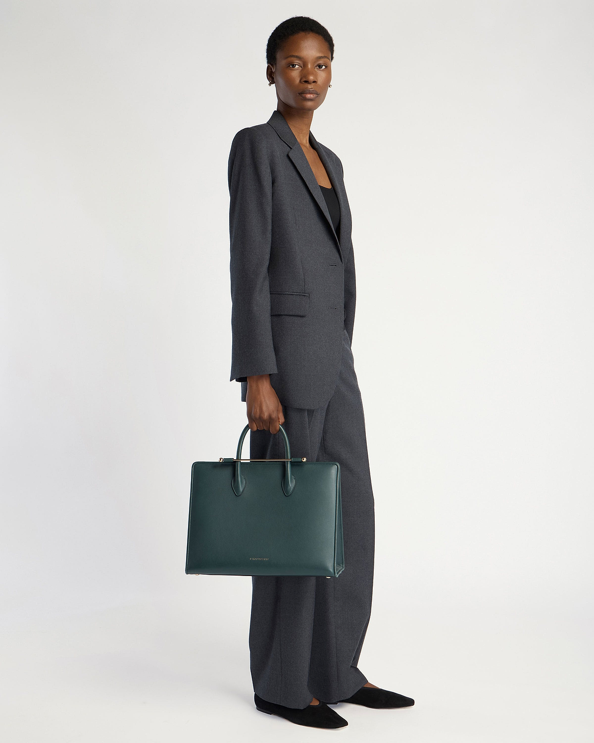 Model holding bottle green leather Strathberry tote bag with top handles.