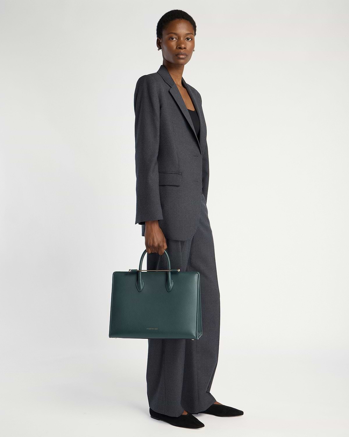 Model holding bottle green leather Strathberry tote bag with top handles.